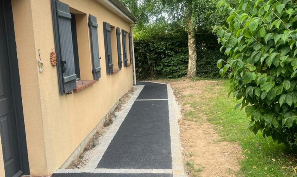 Enrobé pour allée latérale le long d’une maison, bande en enrobé noir bordée de pavés clairs, chemin propre et stable vers le jardin avec arbres et haies.