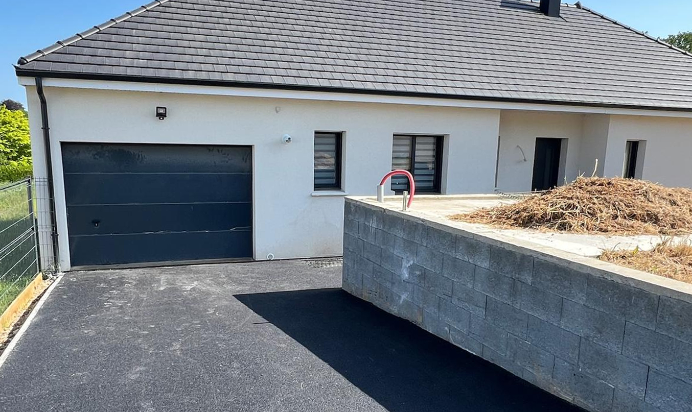 Aménagement extérieur avec chemin en enrobé autour d’une maison, encadrement en pavés clairs, murs en pierre apparente et pelouse sur le côté.