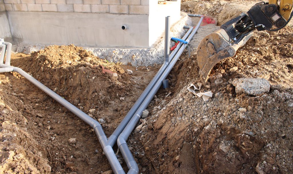 Travaux d’assainissement : pose de canalisations PVC dans une tranchée, avec godet de pelle mécanique à côté.