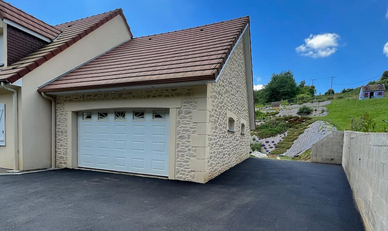 Enrobé et pavés devant garage : vaste aire de stationnement en enrobé noir menant à une porte de garage blanche, bordée d’un muret et d’un talus paysager sous ciel bleu.