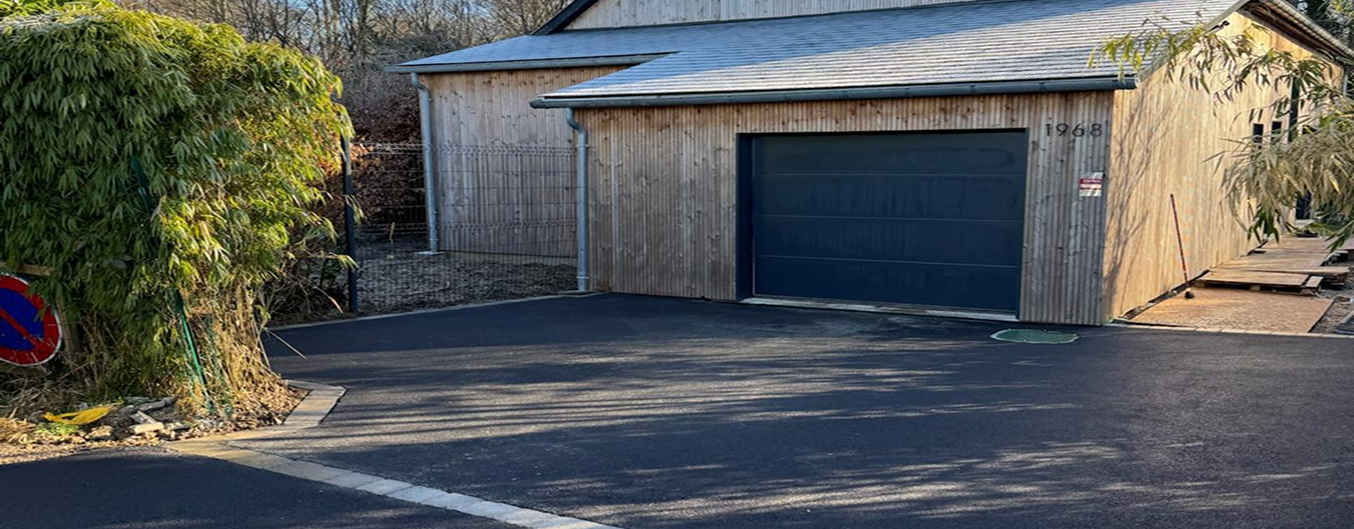 Enrobé et pavés devant garage : grande cour en enrobé noir avec bande de pavés clairs en séparation, accès garage sur bâtiment bardage bois, arbres en arrière-plan.
