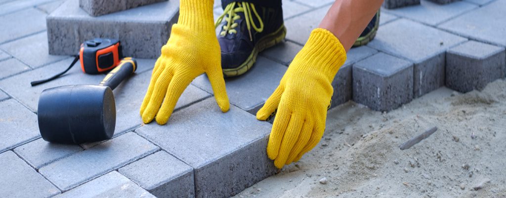 Pose de pavé en béton sur une terrasse, ouvrier agenouillé alignant les pavés, avec sable de réglage et outils visibles.