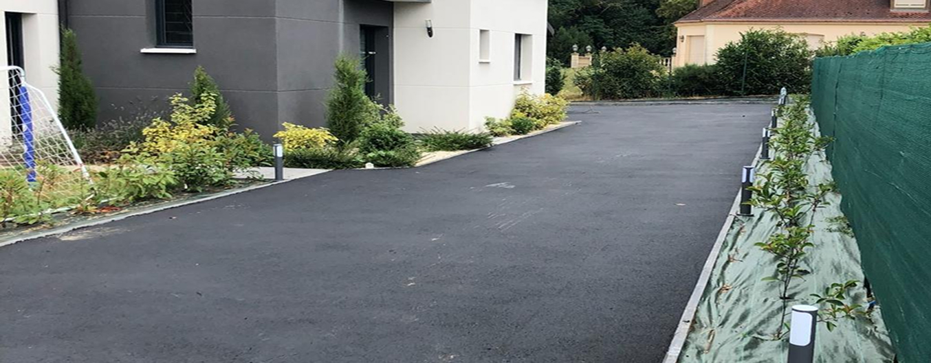 Aménagement extérieur d’une maison contemporaine avec grande allée en enrobé noir, bordures béton et massifs végétalisés le long de la façade.