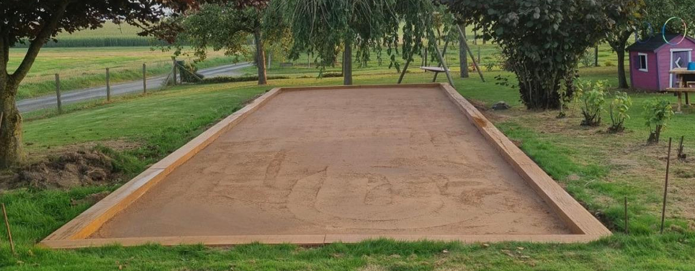 Terrain pétanque maison terminé dans un grand jardin, rectangle de sable compacté bordé de traverses en bois, arbres et campagne en arrière-plan.