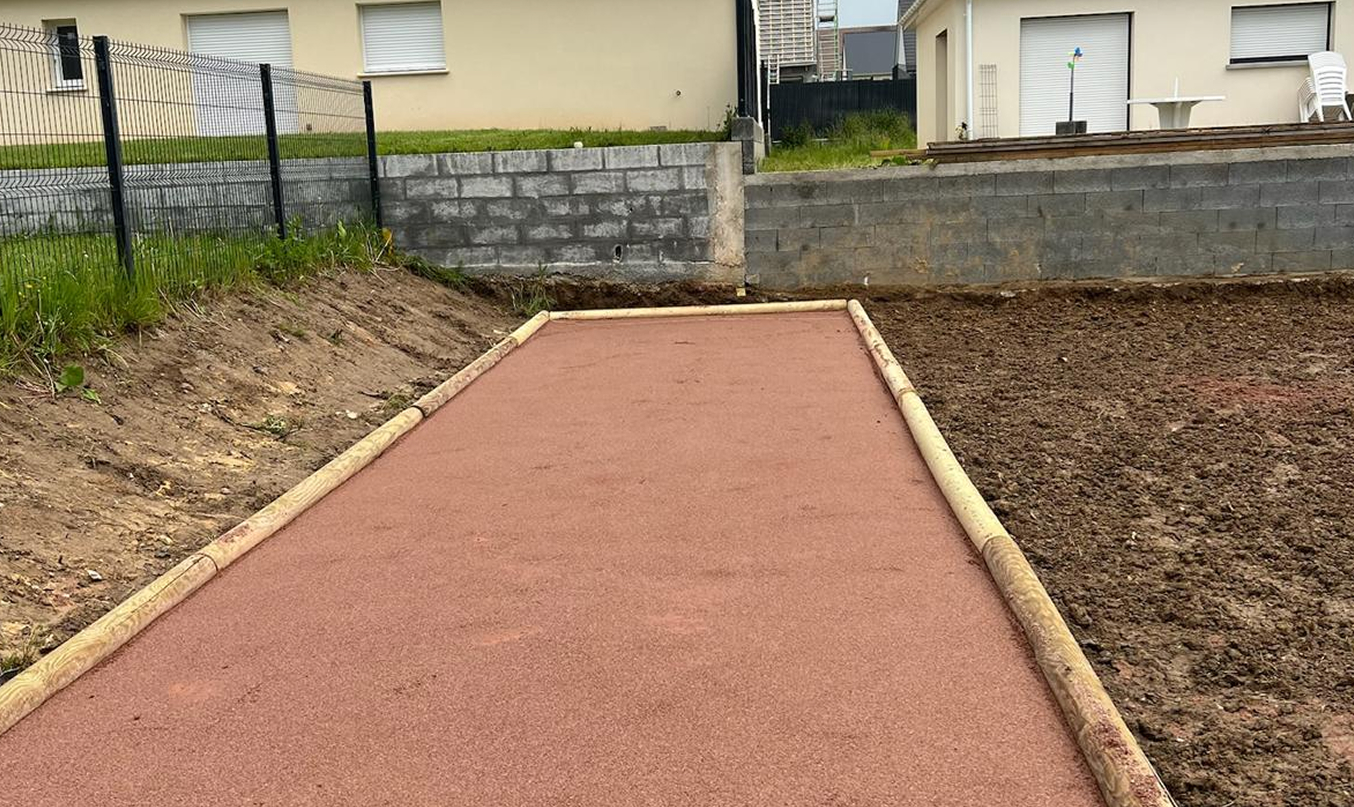 Terrain pétanque maison fraîchement réalisé, bande rectangulaire en gravier rouge bordée de traverses bois, terre préparée autour et maisons au fond.