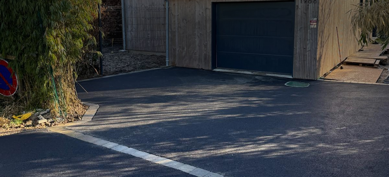 Aménagement extérieur avec chemin en enrobé autour d’une maison, encadrement en pavés clairs, murs en pierre apparente et pelouse sur le côté.
