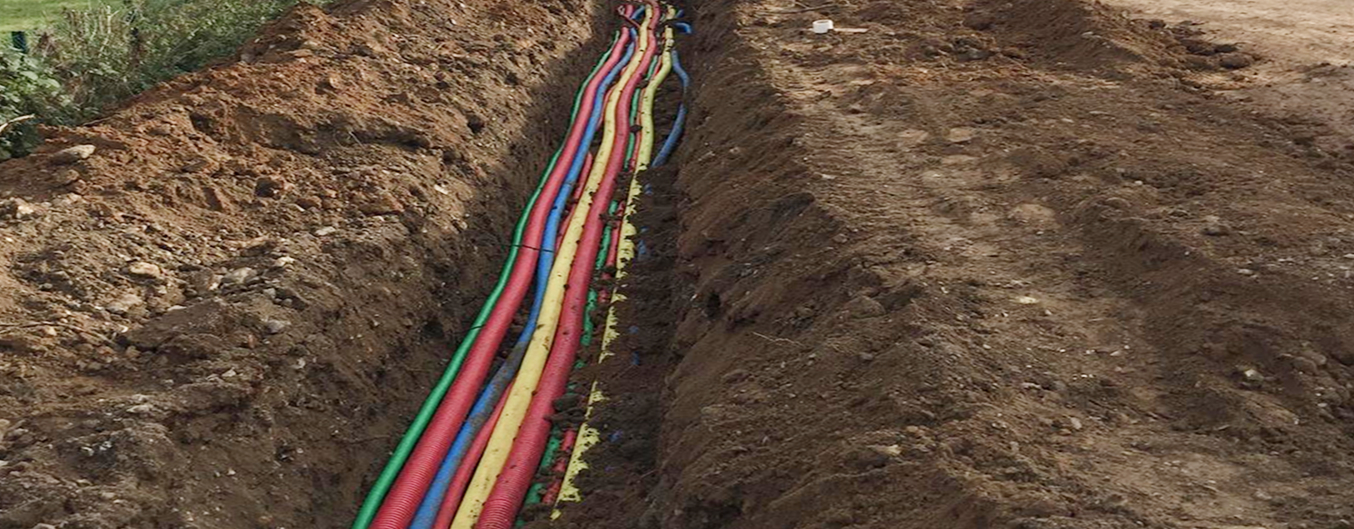 Tranchée technique longue avec réseaux enterrés (gaines multicolores) et pelleteuse au second plan sur chantier extérieur.