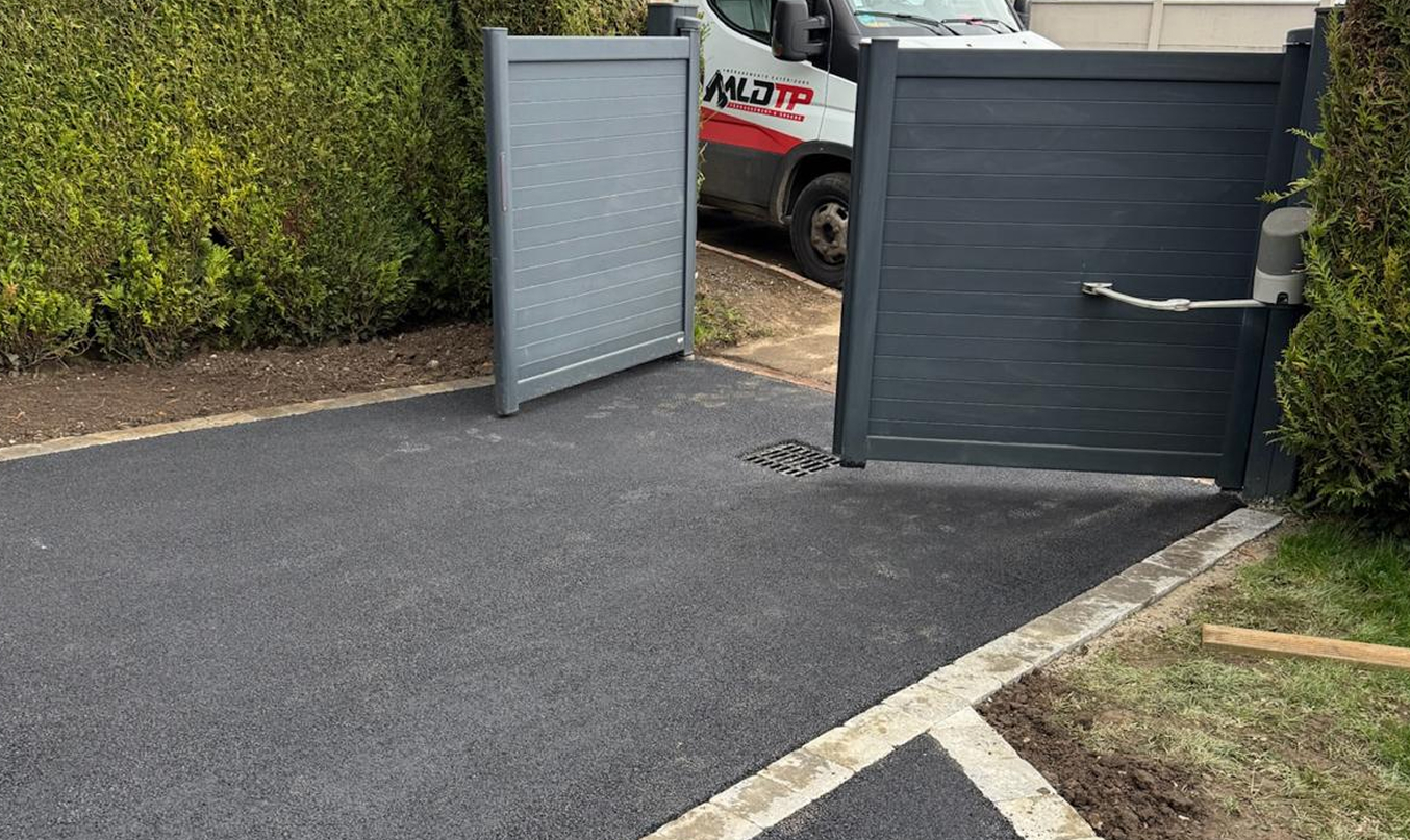 Enrobé pour cour avec portail battant gris ouvert, entrée asphaltée neuve et bordures en pavés clairs au milieu des haies.