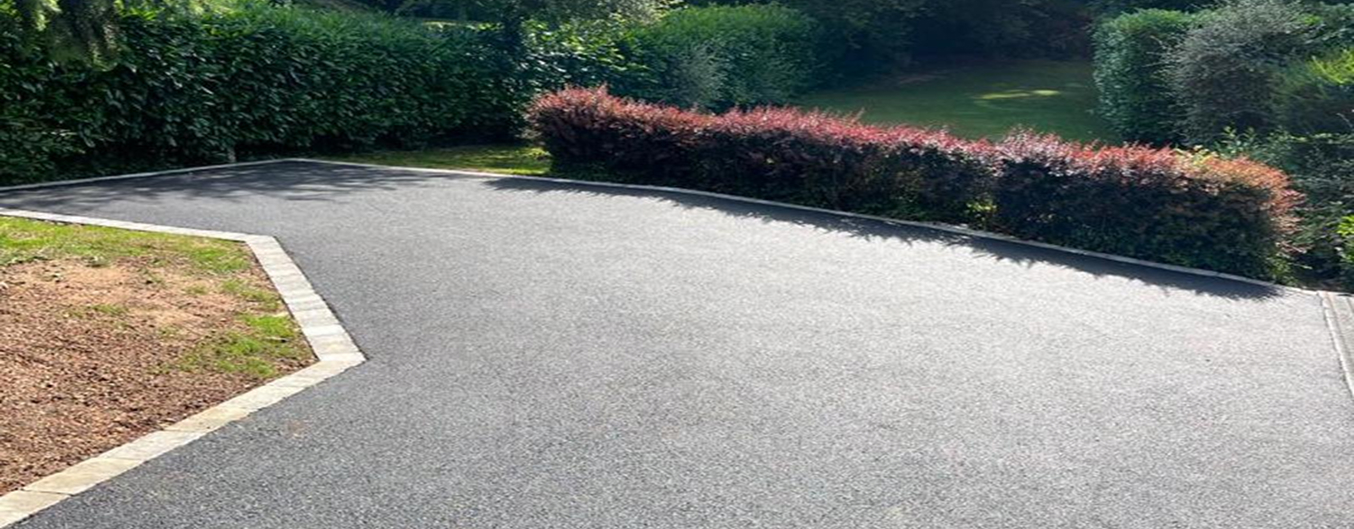 Enrobé pour cour dans un jardin arboré, grande surface asphaltée lisse bordée de pavés, avec massifs et haies en arrière-plan.